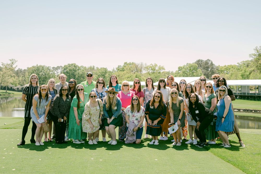 Large group of women posing outdoors on a sunny day near a pond at a golf course event.