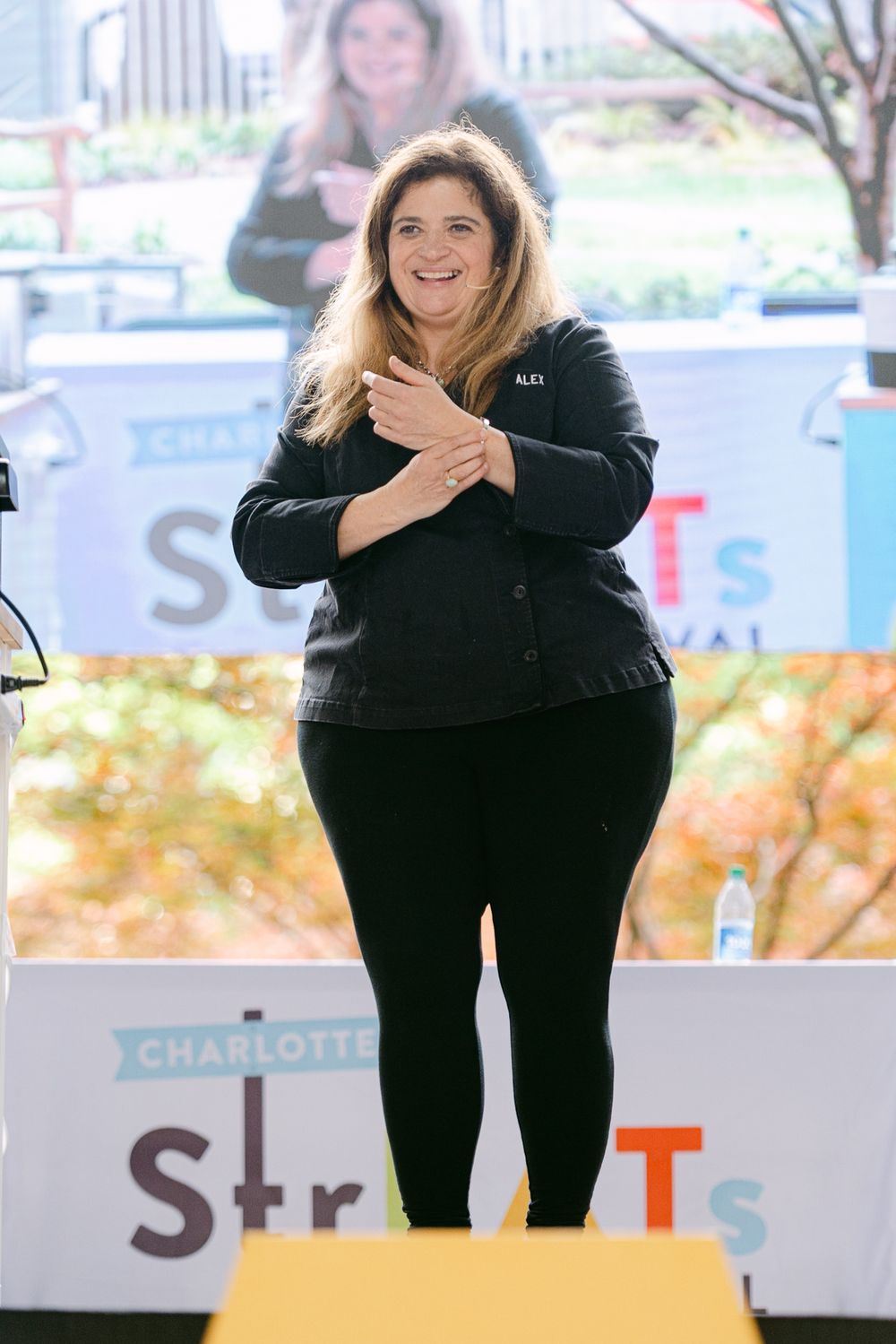 Woman in black jacket smiles on stage at Charlotte Streets Festival outdoor event.