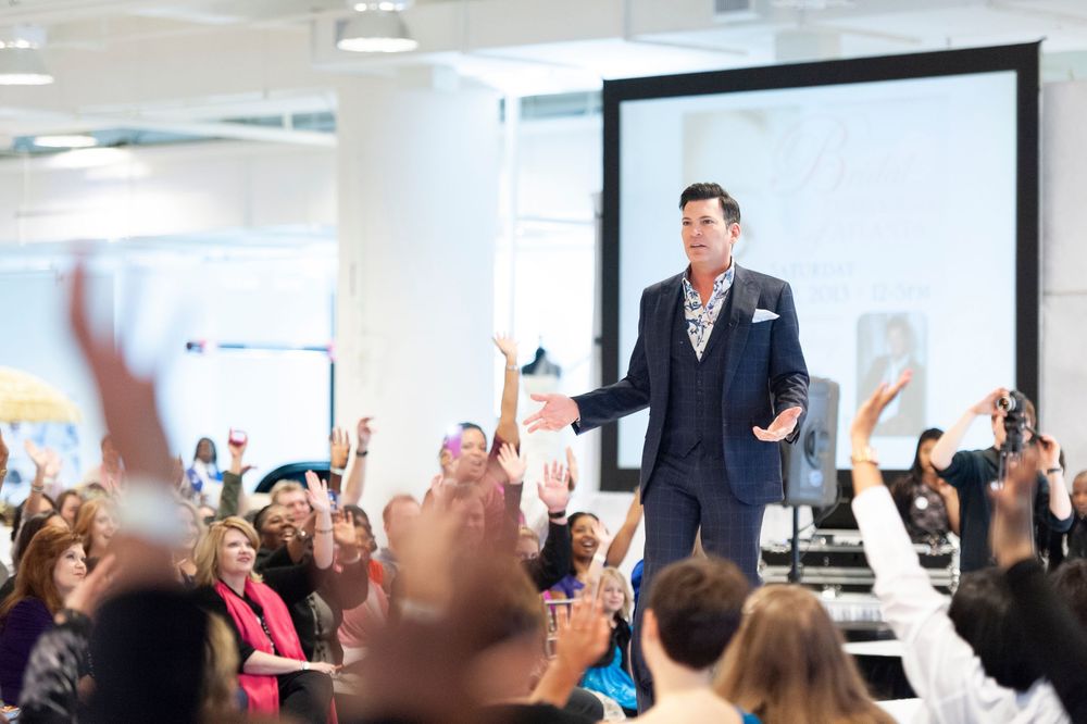 Man in navy suit speaks on stage to enthusiastic crowd with raised hands at a bright indoor event.