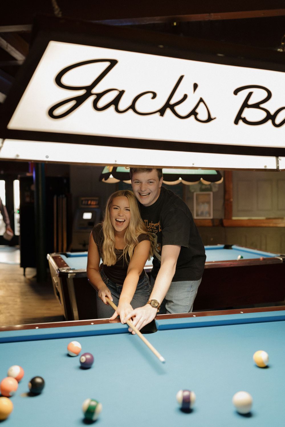 Couple laughing and playing pool at Jack's Bar, posing with cue sticks over a billiard table.