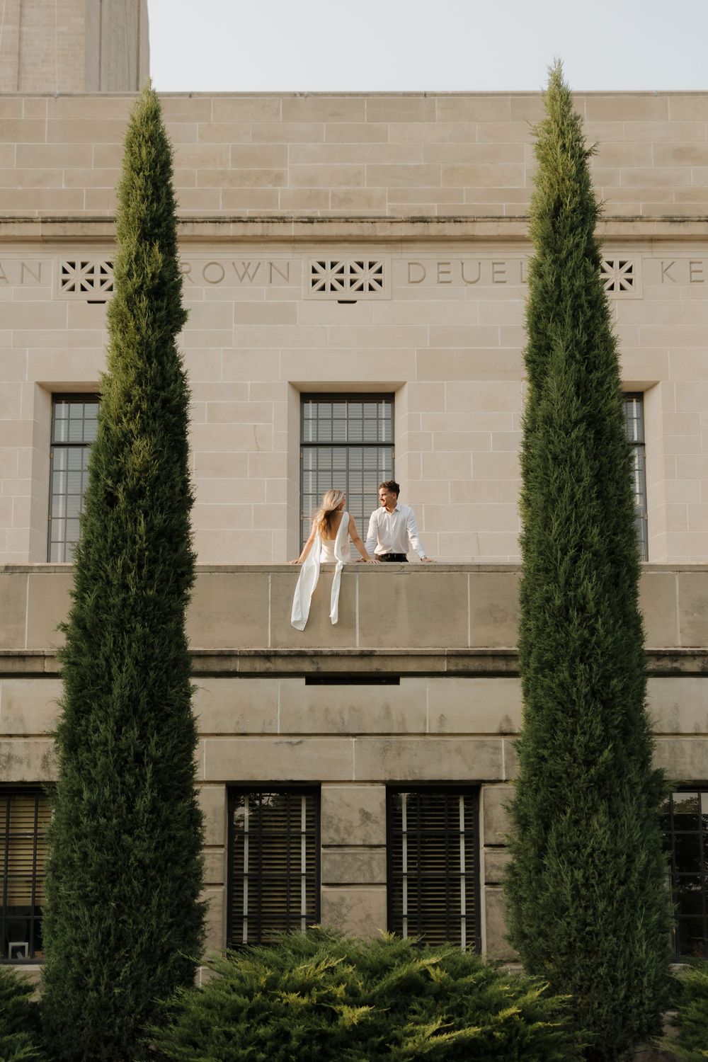 Couple in white attire standing on a stone ledge of a classical building flanked by tall cypress trees.