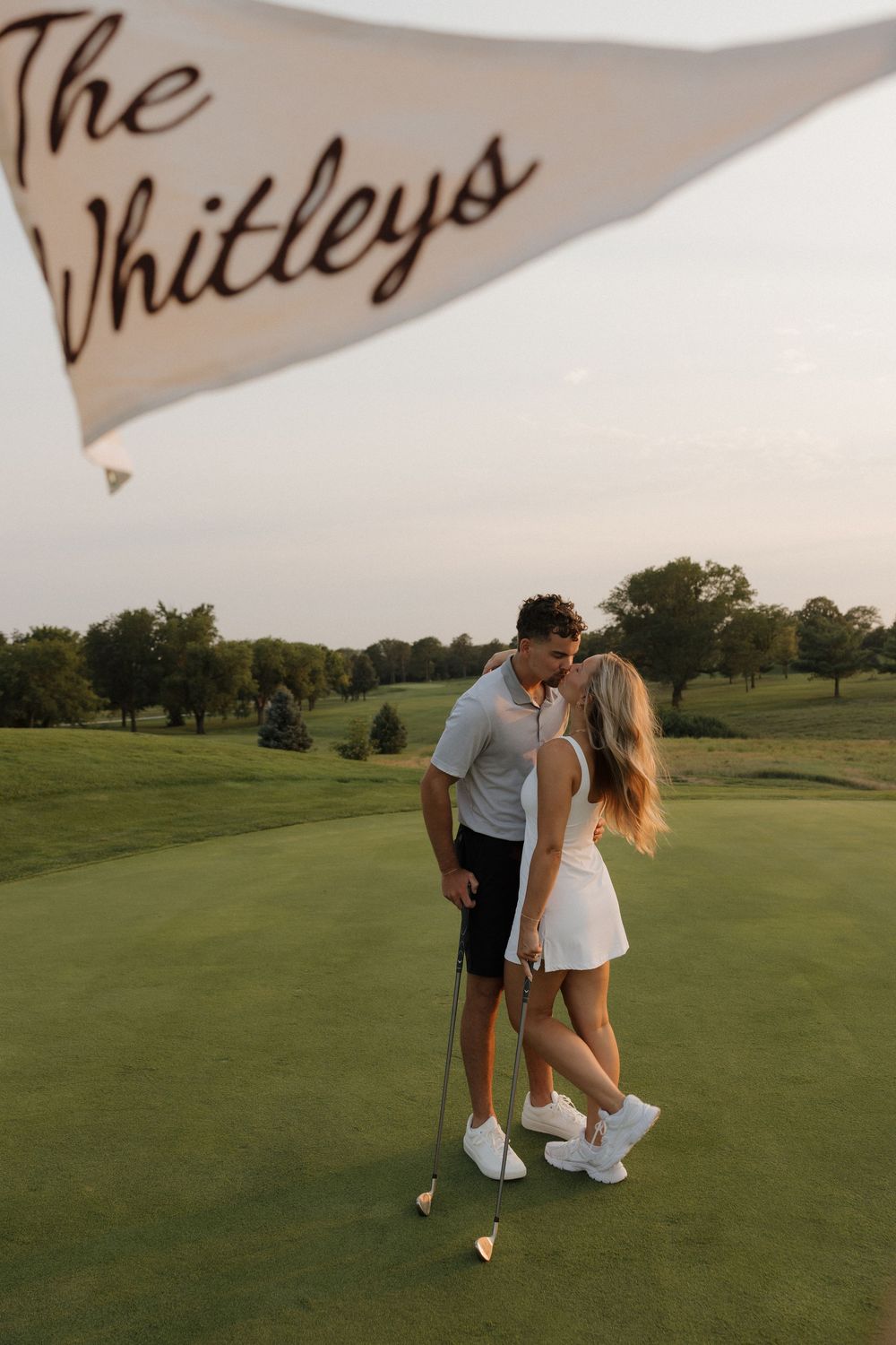 Couple sharing a romantic moment on a golf course holding a flag reading The Whitleys during golden hour.