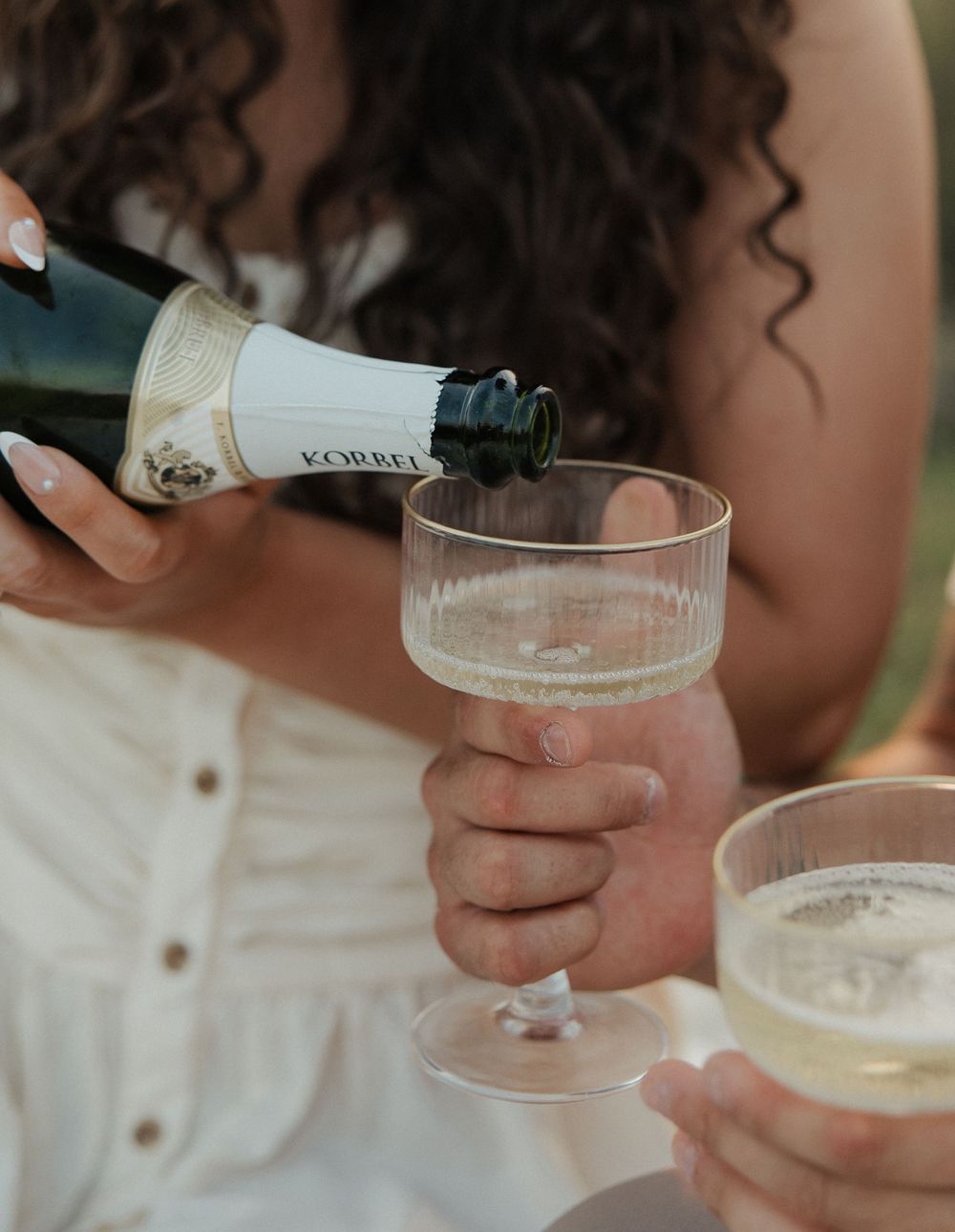 Hands pouring champagne into elegant coupe glasses during an outdoor celebration.