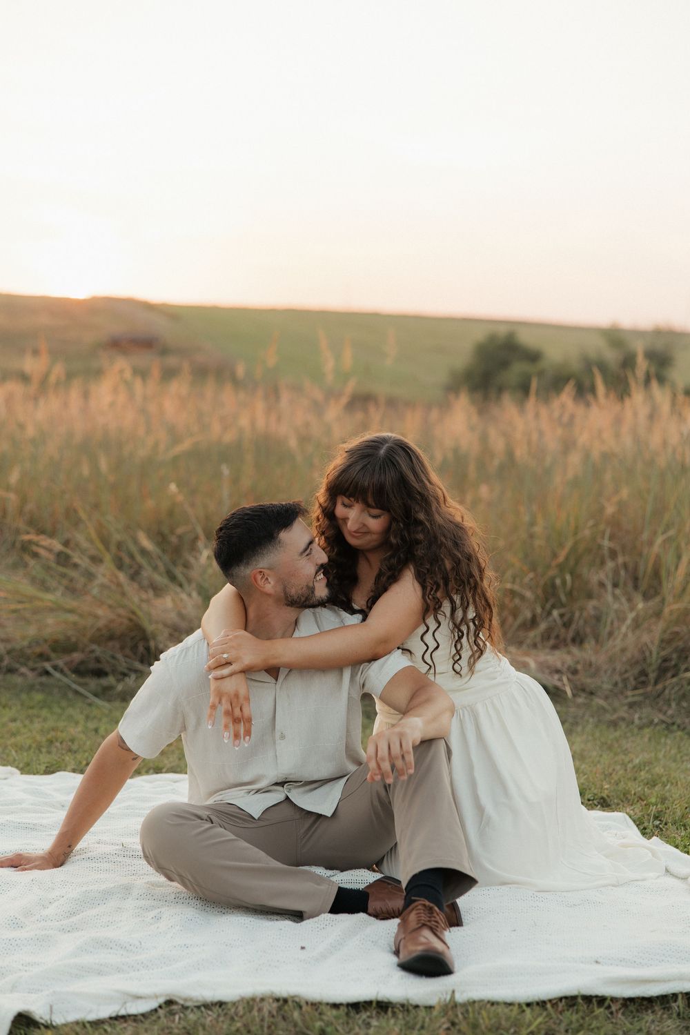 A couple sits intimately on a white blanket in a grassy field during golden hour.
