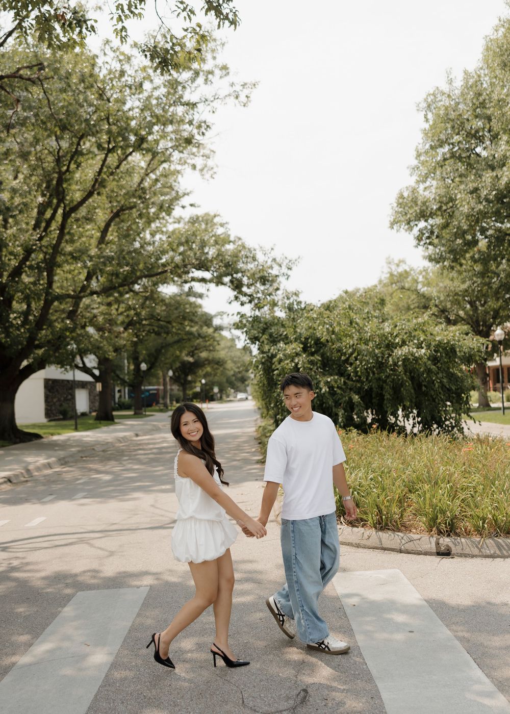 Couple holding hands and crossing a tree-lined street, woman in white dress and man in white tee with jeans.