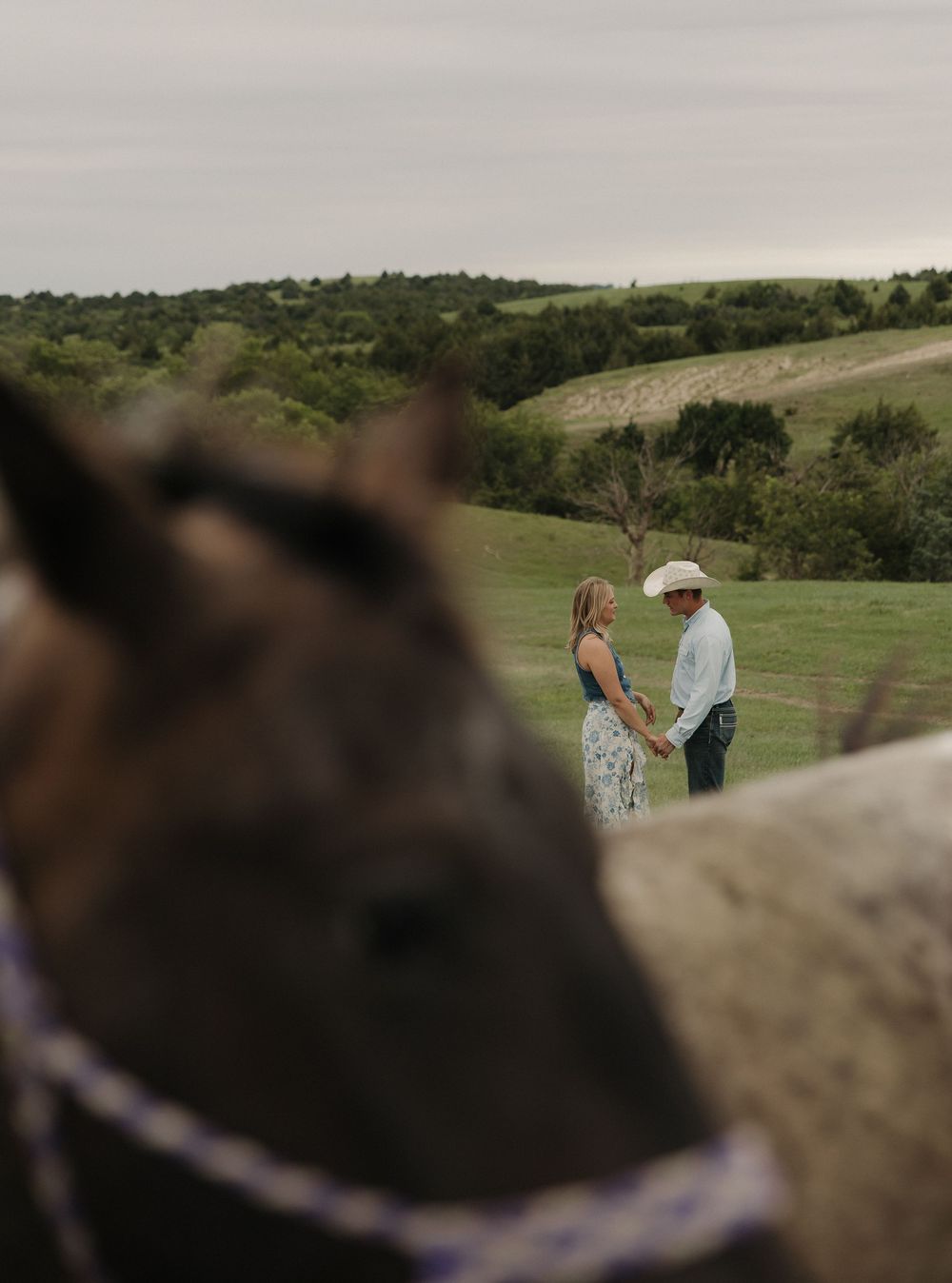 A couple shares a romantic moment on a grassy hillside with rolling green hills in the background during golden hour.