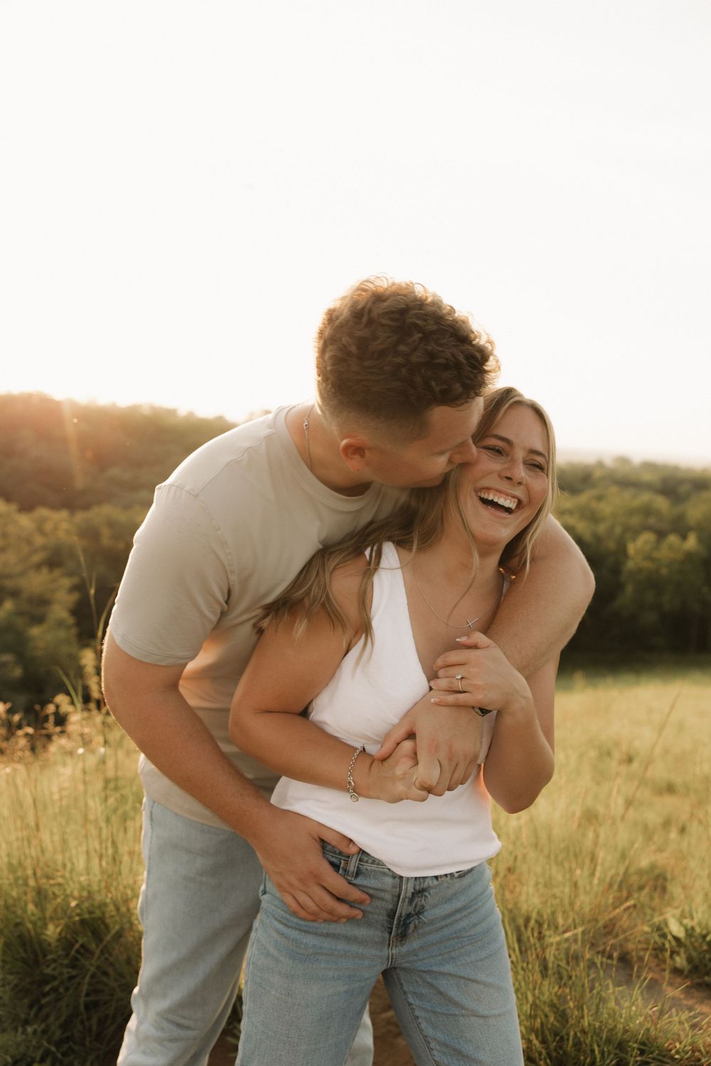 A couple embraces and laughs together in a sunlit meadow during golden hour.