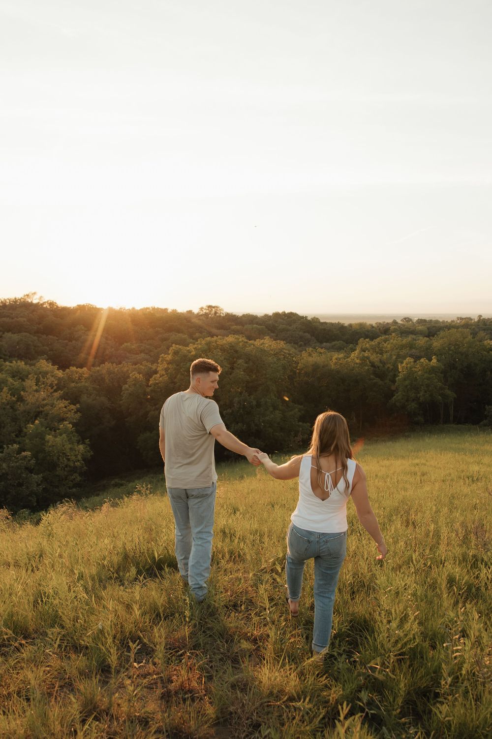 Couple walking hand in hand through a golden meadow at sunset with trees in the background.