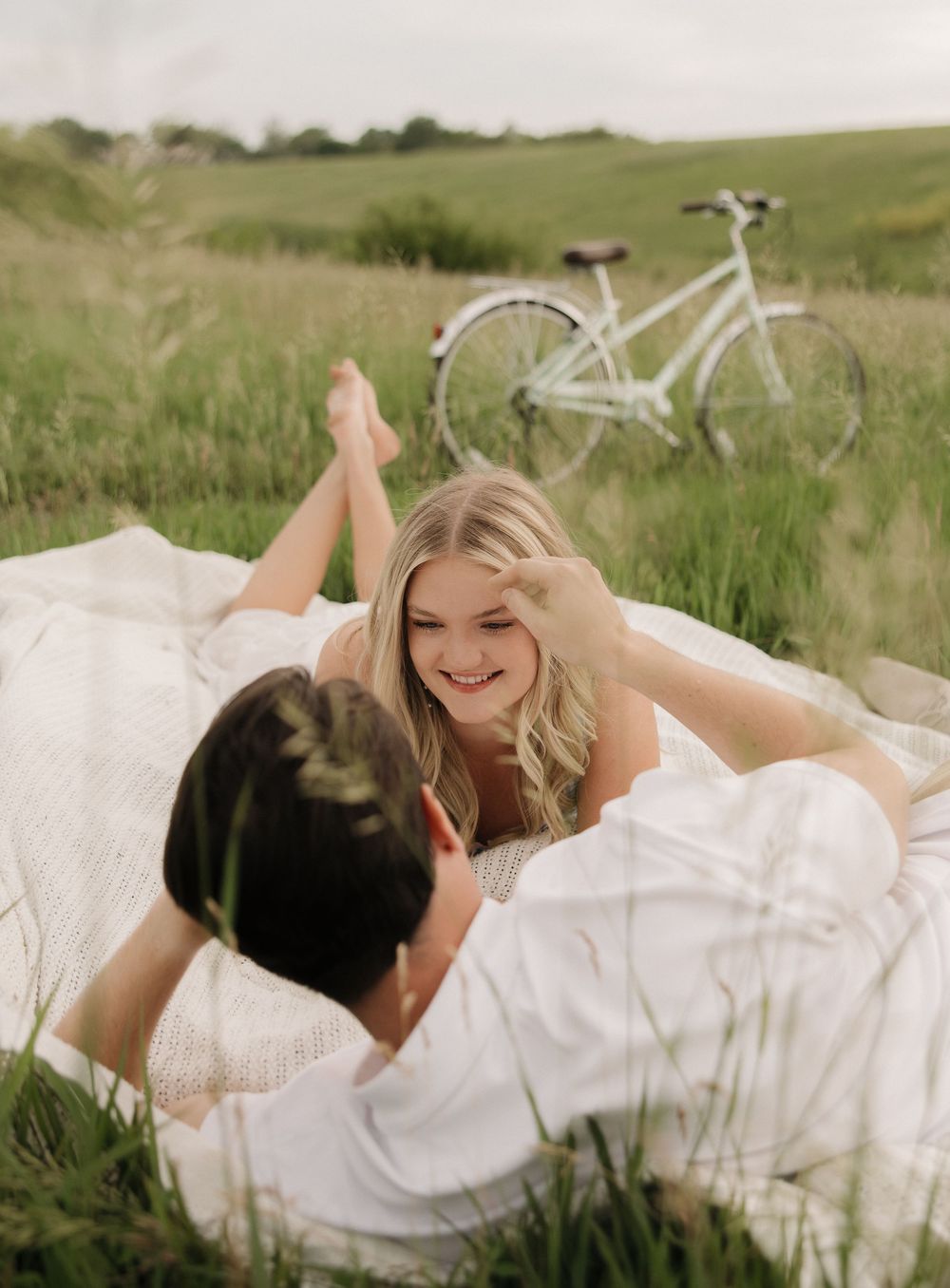 A romantic couple lying in tall grass wearing white outfits while sharing intimate moments in a natural outdoor setting.