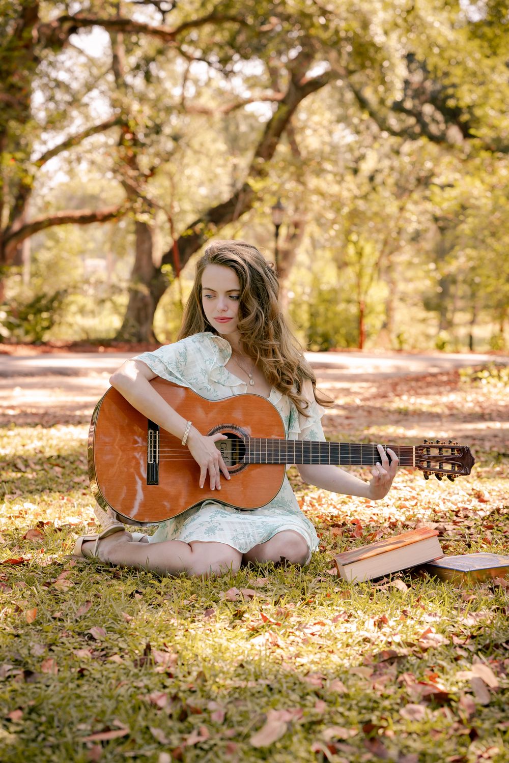 A person in a white dress sits on autumn leaves while playing an acoustic guitar in a sun-dappled outdoor setting.