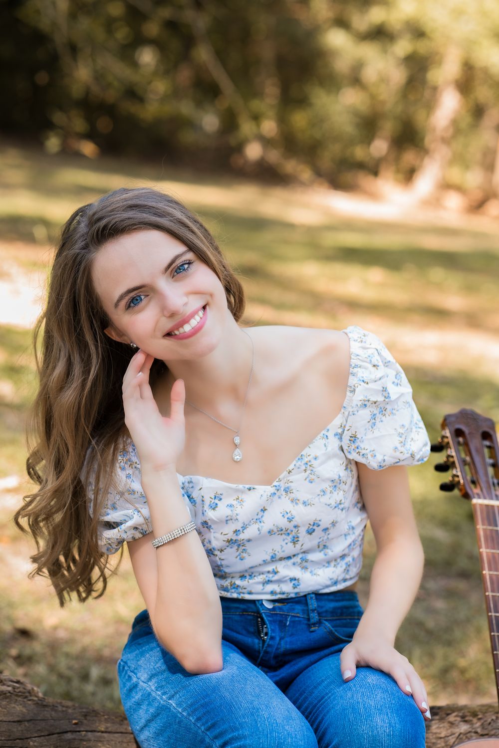 A person wearing a white floral top and blue jeans sits casually outdoors in a natural setting with sunlit trees.