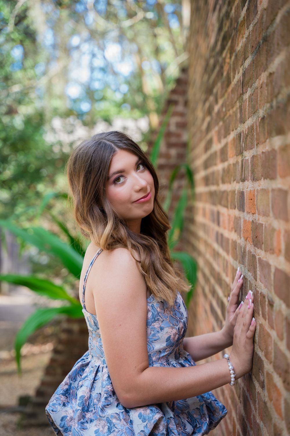 Person in a floral blue dress poses against a rustic brick wall in an outdoor garden setting.