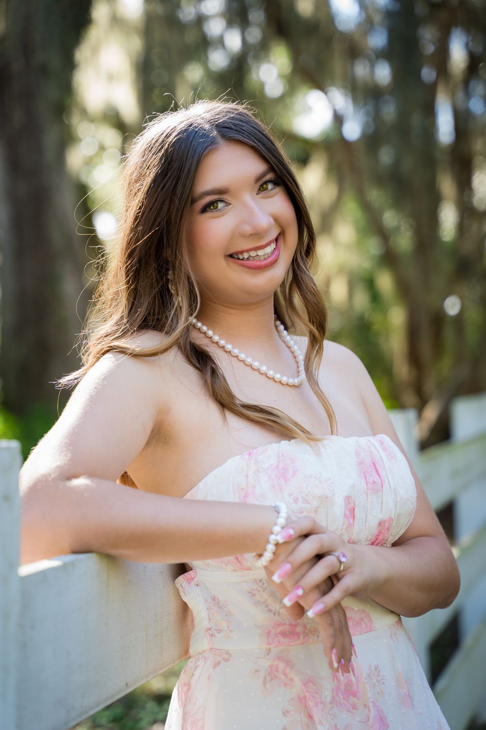 A person in a white dress with pink accents poses playfully in a wooded outdoor setting wearing a pearl necklace.