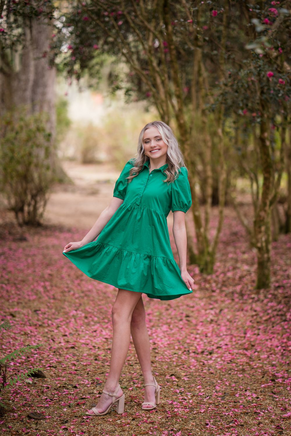 A vibrant emerald green dress twirls gracefully among fallen pink flower petals in a wooded outdoor setting.