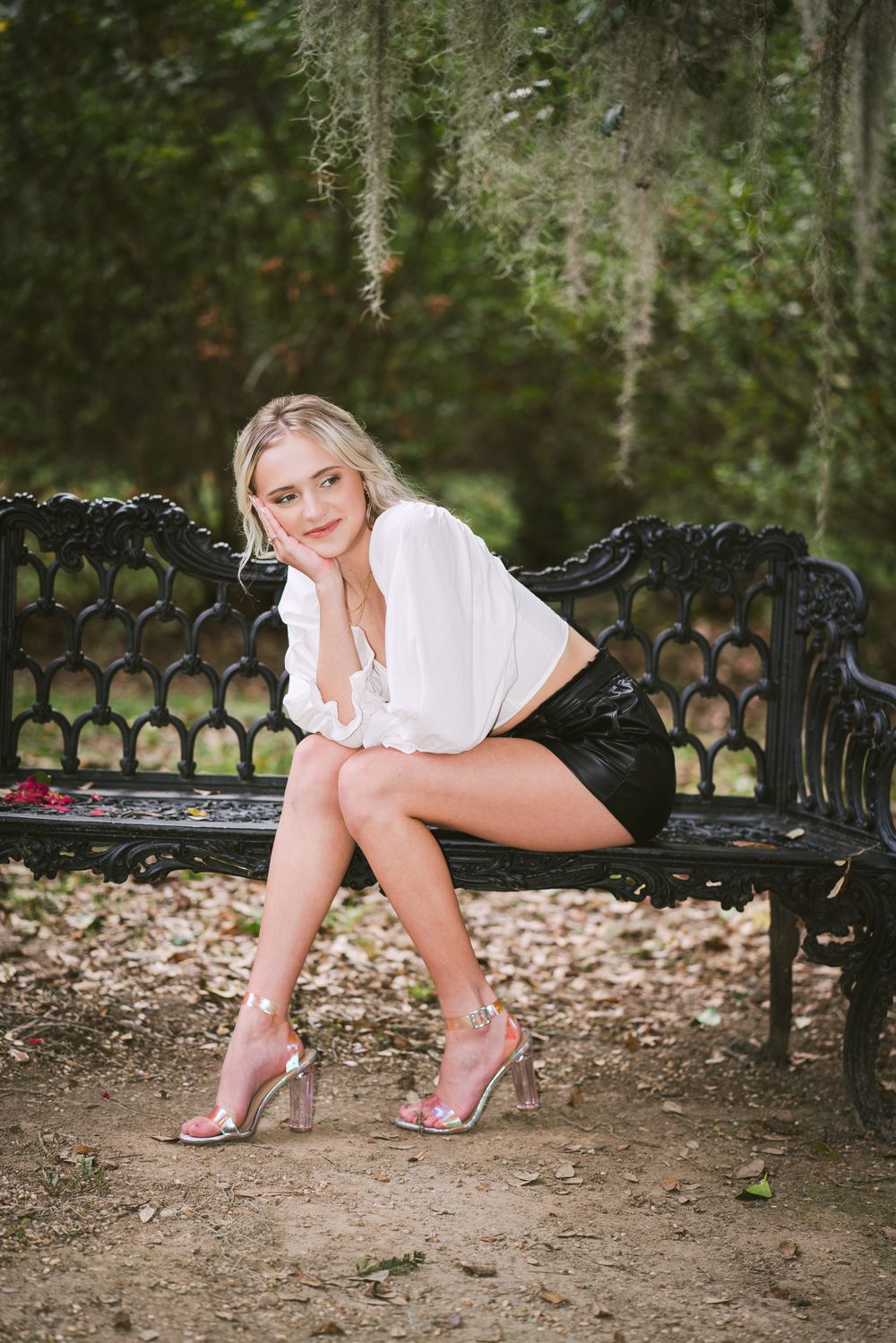 A person in a white top and black shorts sits elegantly on an ornate black metal garden bench under Spanish moss.