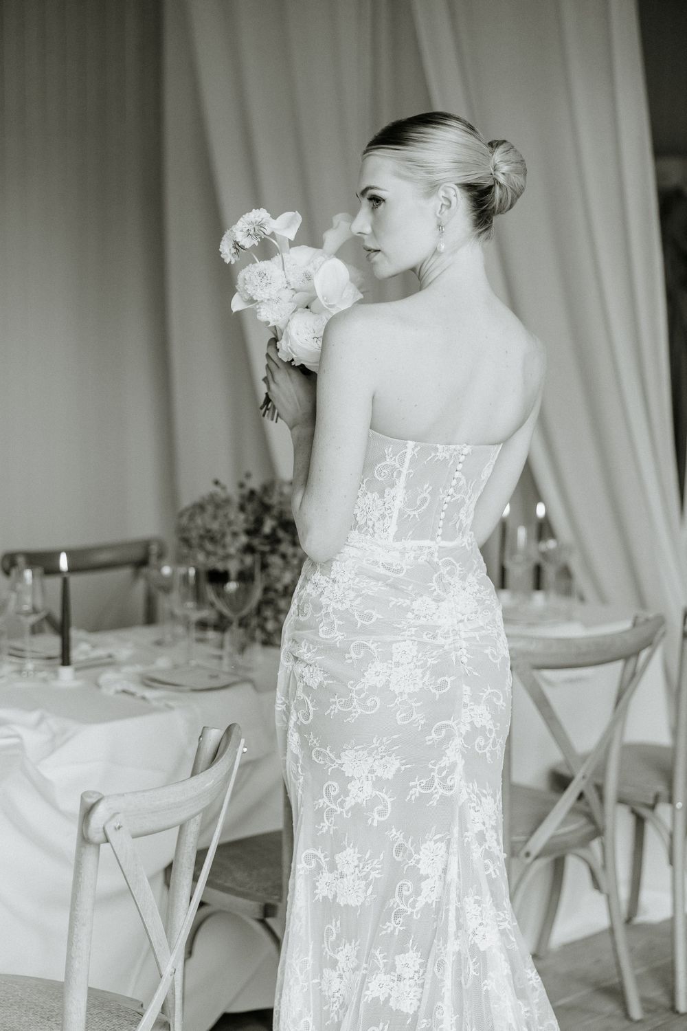 Black and white photograph of bride in fitted lace wedding gown holding bouquet at elegant reception venue.