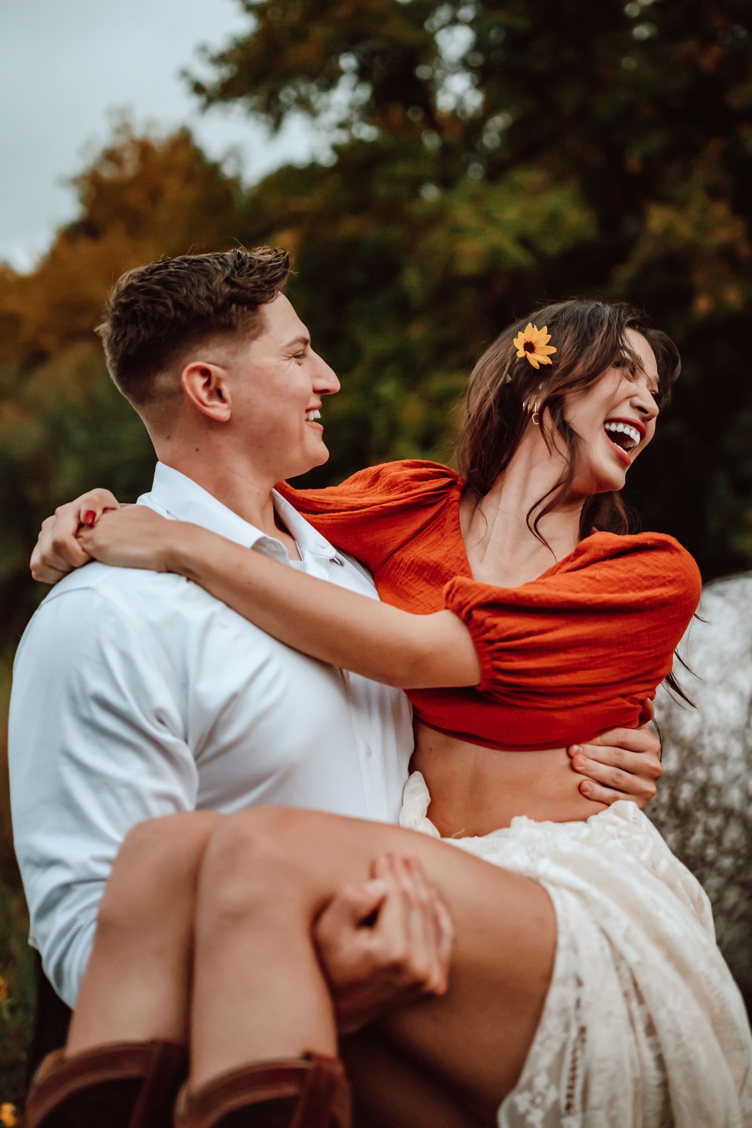 A joyful couple embraces outdoors during autumn, with her wearing an orange top and white skirt against fall foliage.