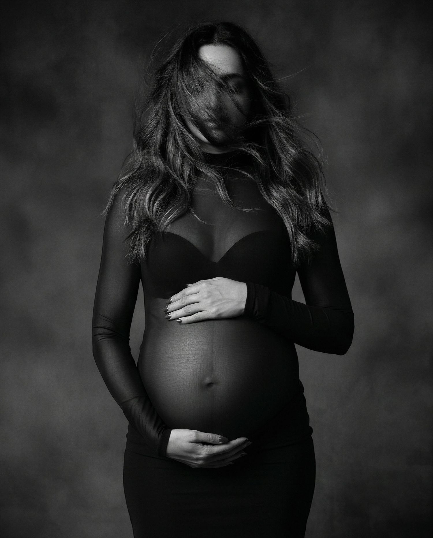 Black and white maternity photo of pregnant woman in black dress cradling baby bump in professional studio portrait.