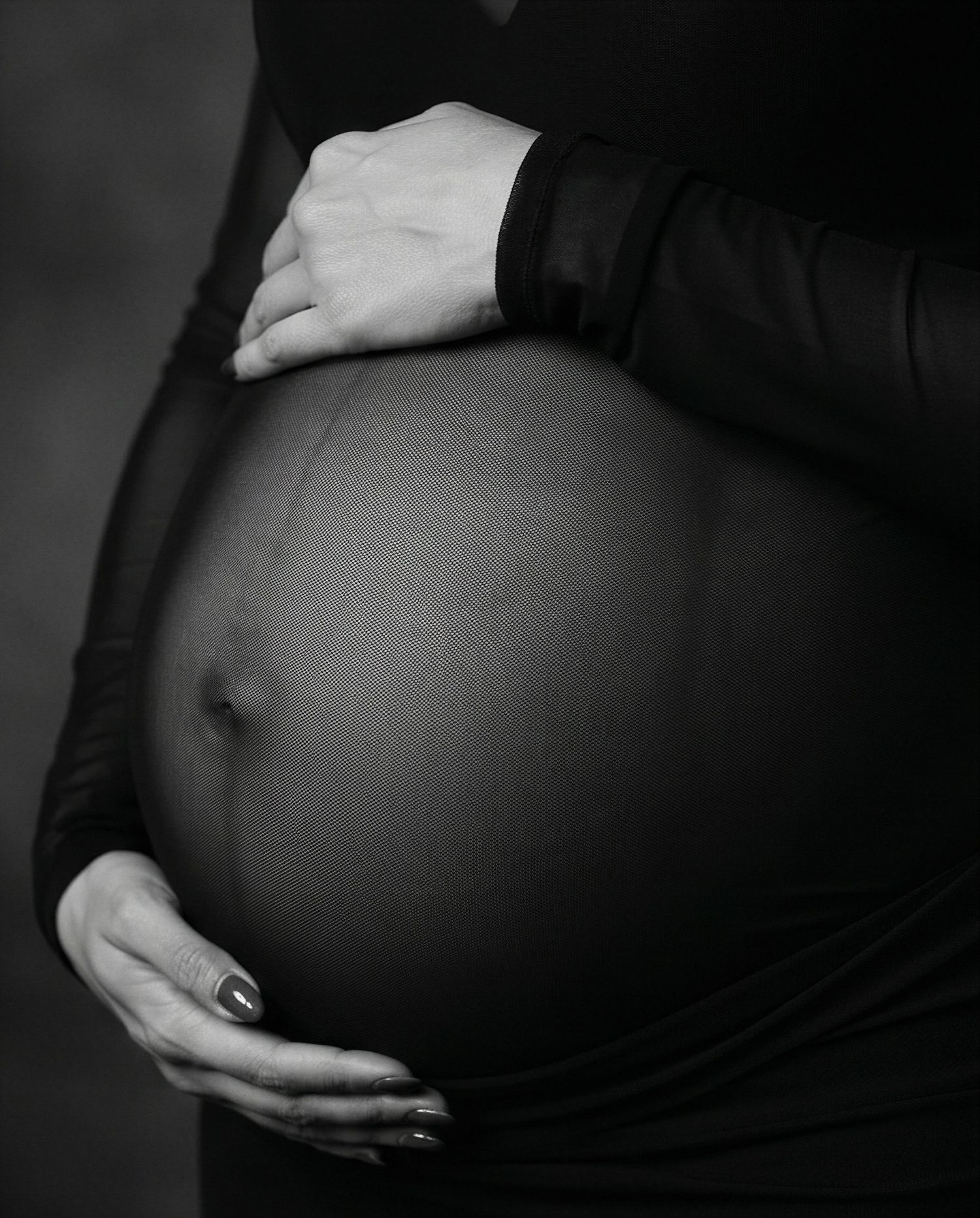 Black and white maternity photo of pregnant woman in black dress cradling her bare baby bump with both hands.