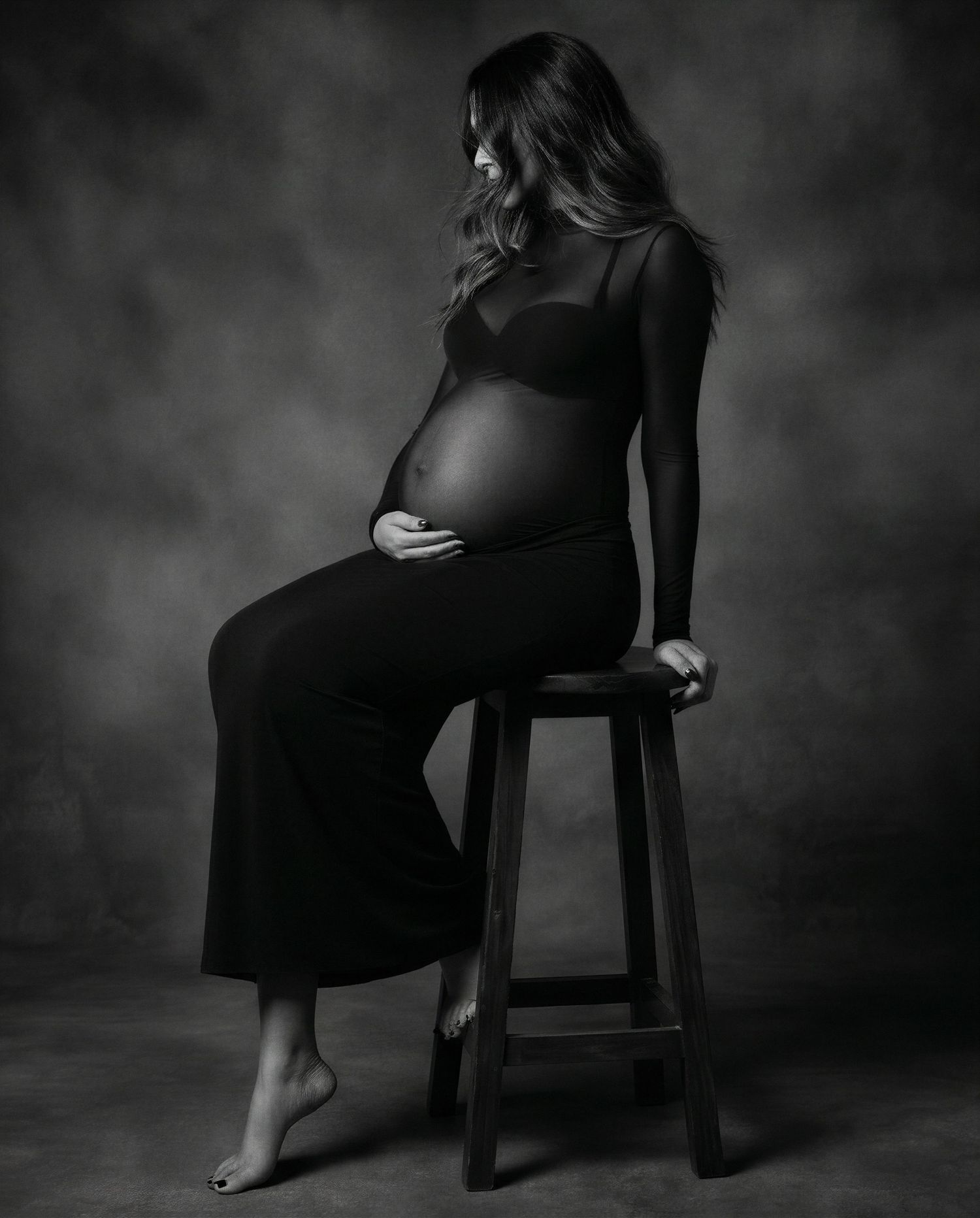Black and white maternity photo of a pregnant woman sitting on a stool, cradling her baby bump in a studio setting.