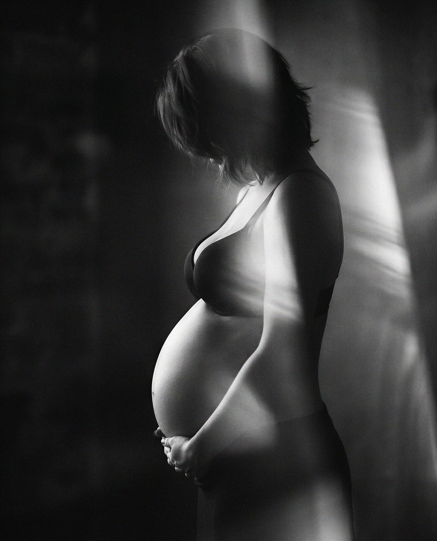 Black and white maternity photo of pregnant woman cradling baby bump in dramatic side lighting.