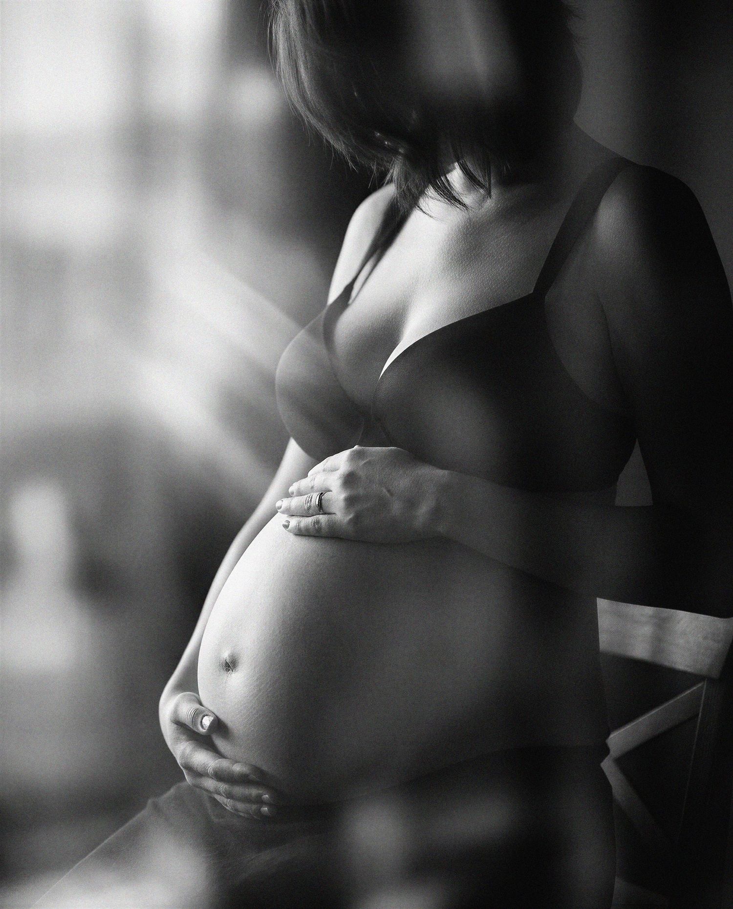 Black and white maternity photo of pregnant woman in bra cradling baby bump near window light.