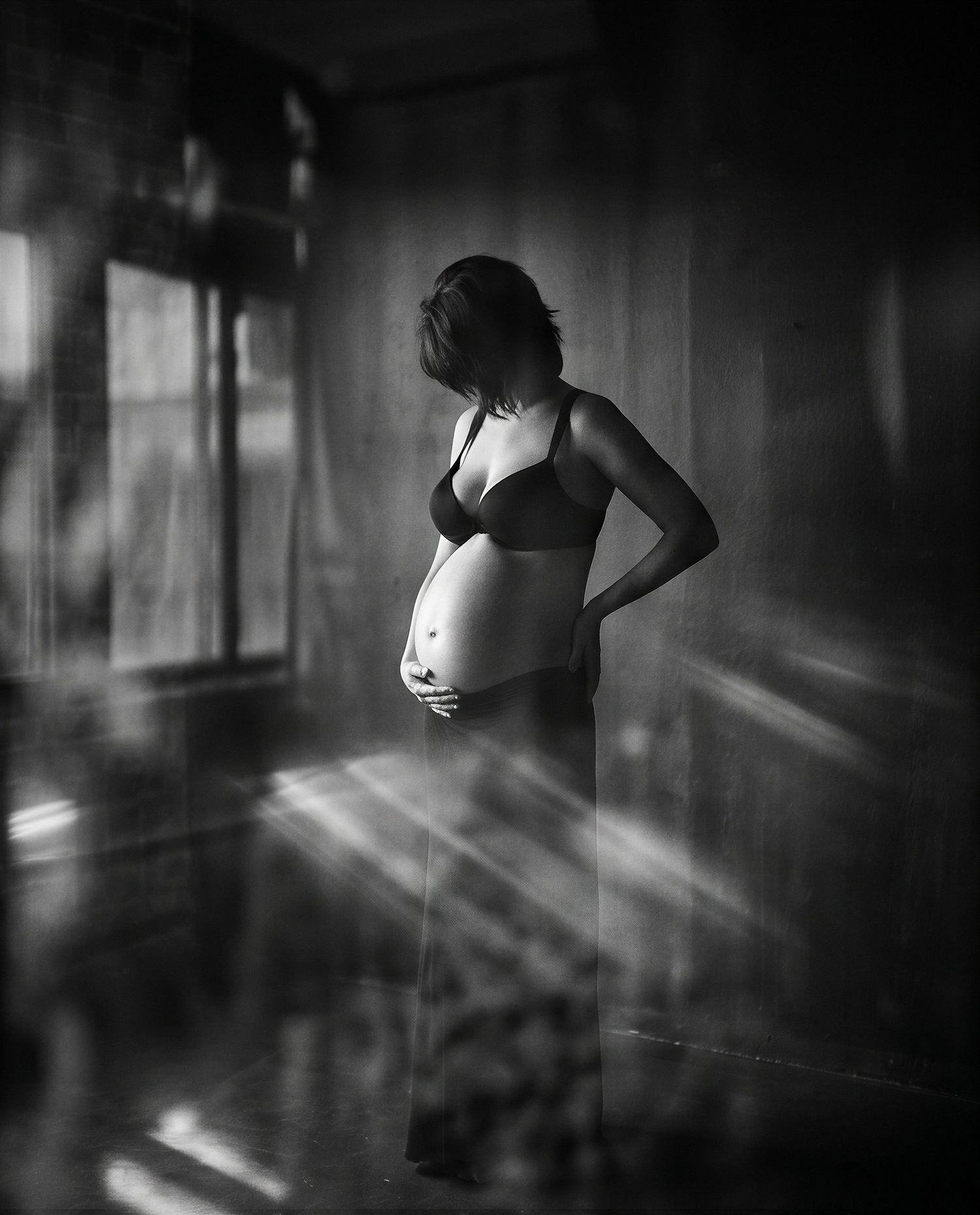 Black and white photo of a pregnant woman in lingerie standing in sunlight streaming through a window.