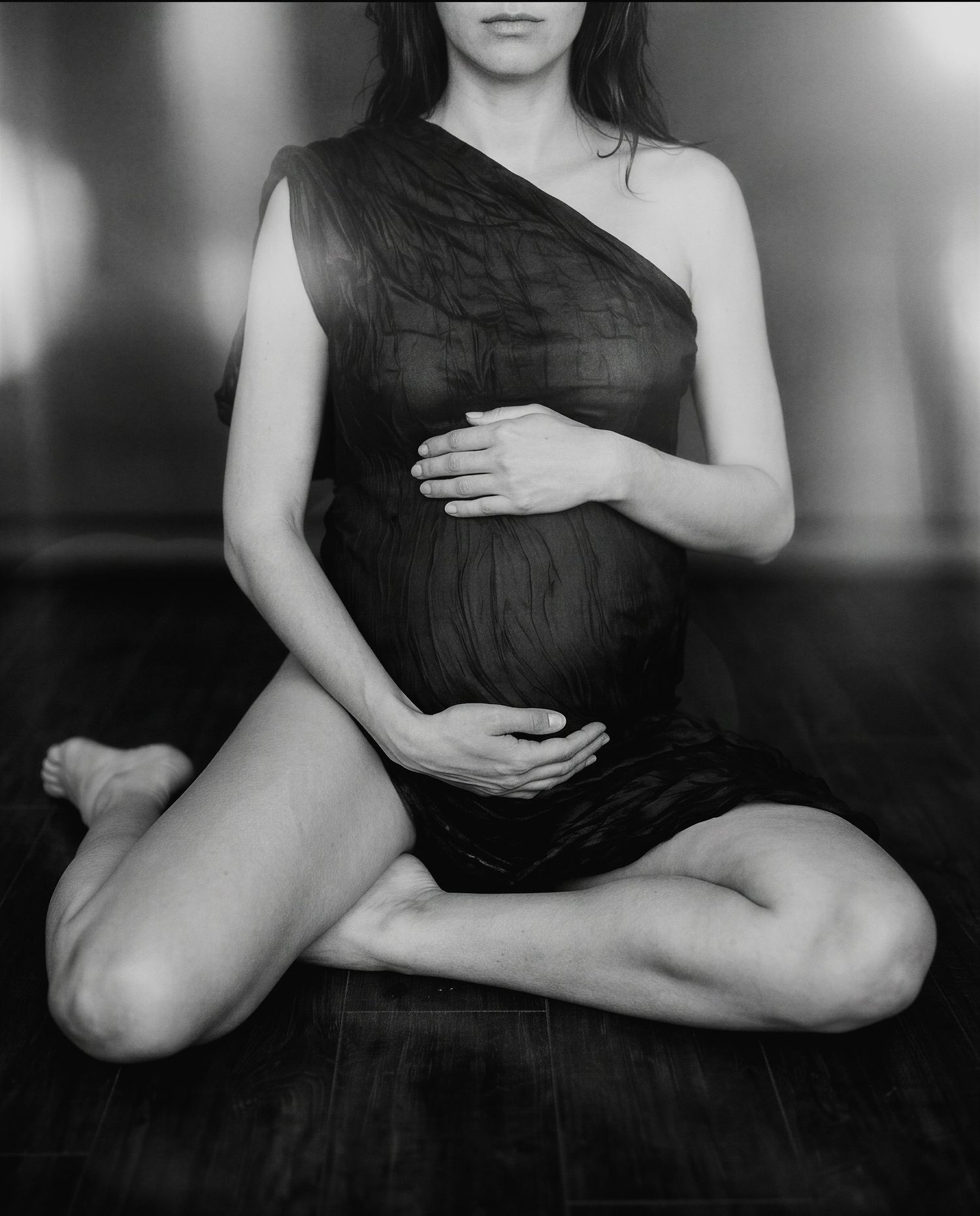 Black and white maternity photo of pregnant woman in sheer dress sitting cross-legged on floor cradling her baby bump.