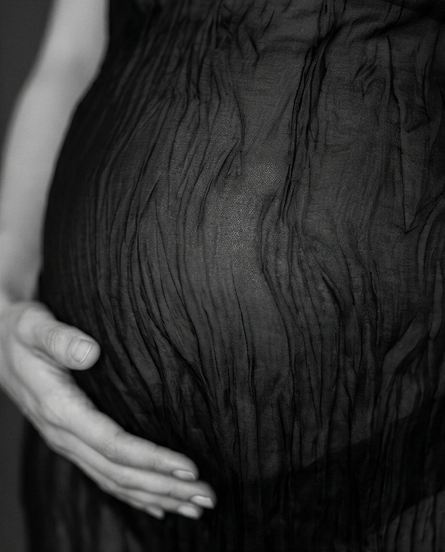 Black and white close-up of a pregnant woman cradling her baby bump through a textured dark fabric dress.