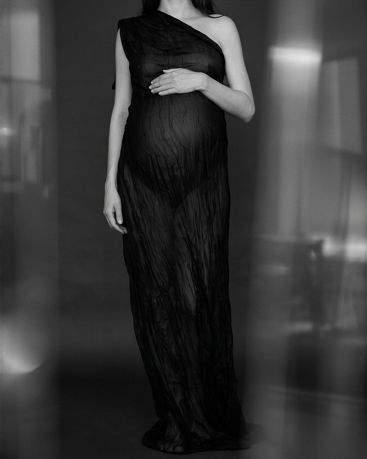 Black and white maternity photo of pregnant woman in elegant one-shoulder black gown, cradling baby bump.