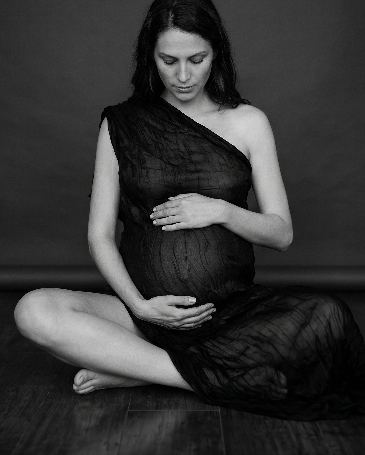 Black and white maternity photo of a pregnant woman in a sheer dress sitting cross-legged, cradling her baby bump.