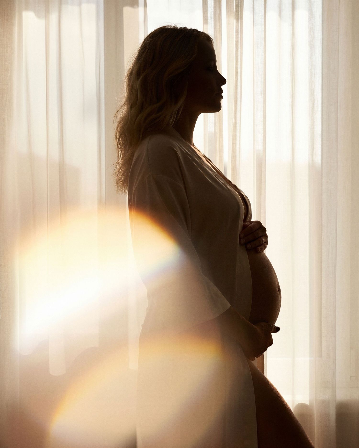 Silhouette of pregnant woman in white robe standing by sheer curtains with golden sunlight and lens flare.