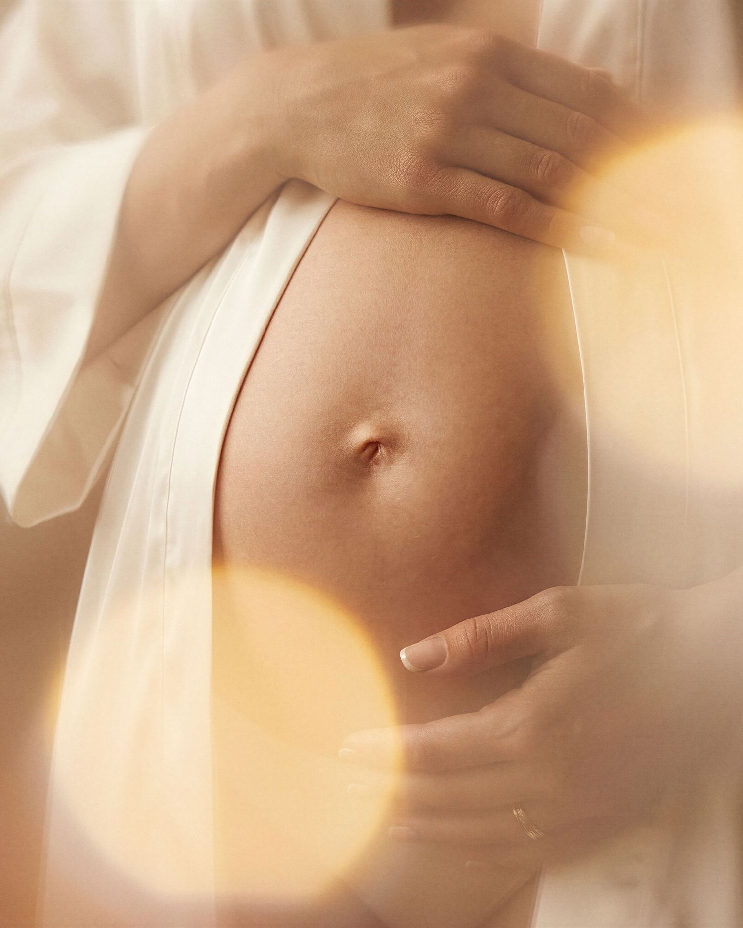 Close-up of a pregnant woman cradling her bare baby bump, wearing a white robe in soft warm glowing light.