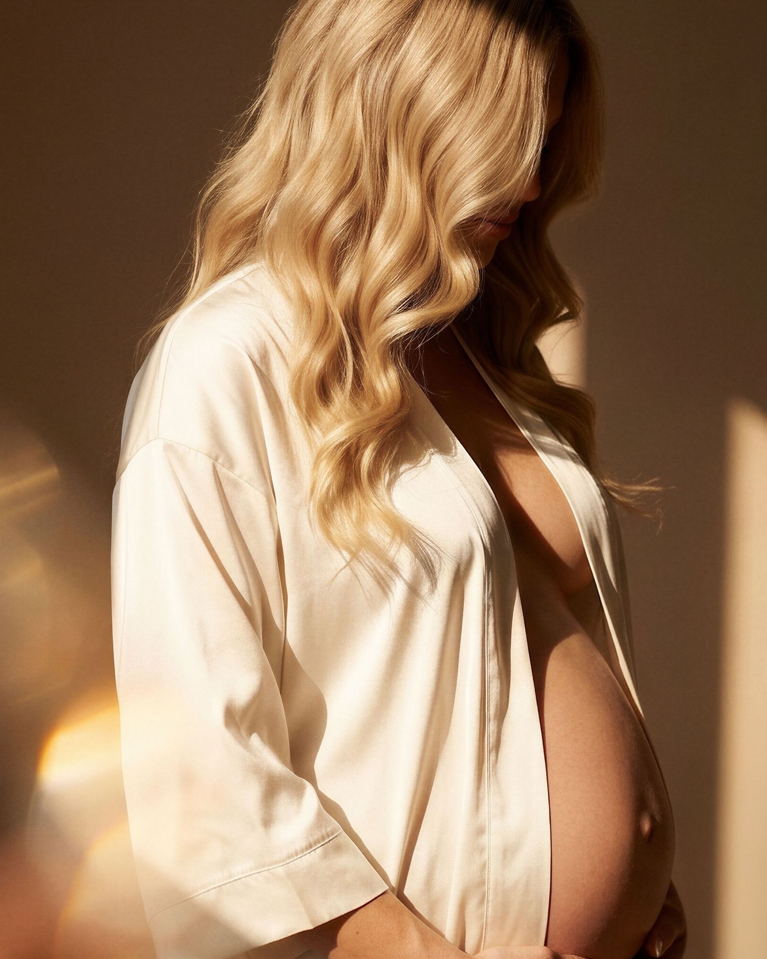 Pregnant woman in silk robe with blonde wavy hair posing sideways in warm golden light.