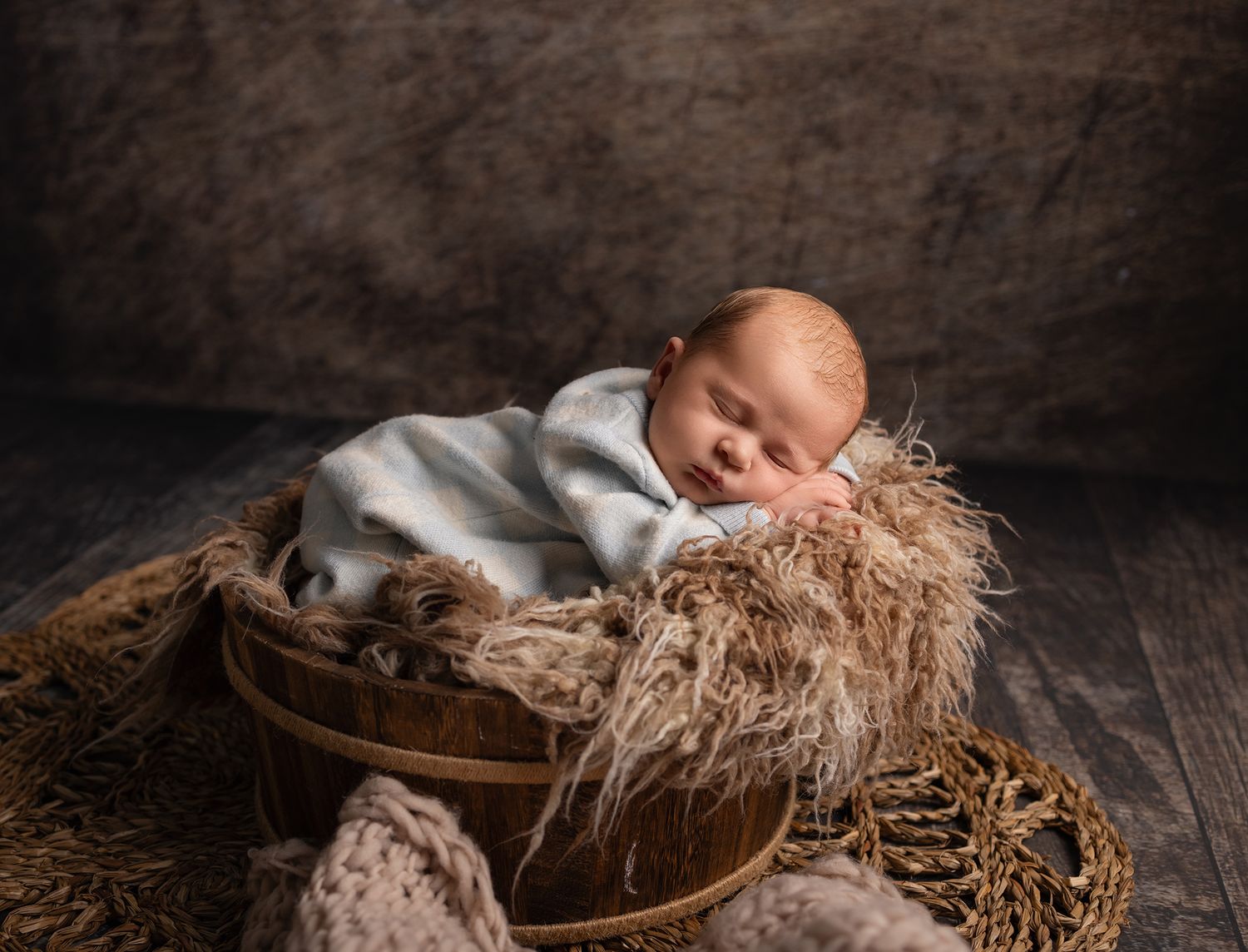 A sleeping newborn baby lies peacefully on soft brown fur in a rustic wooden bowl against a dark backdrop.