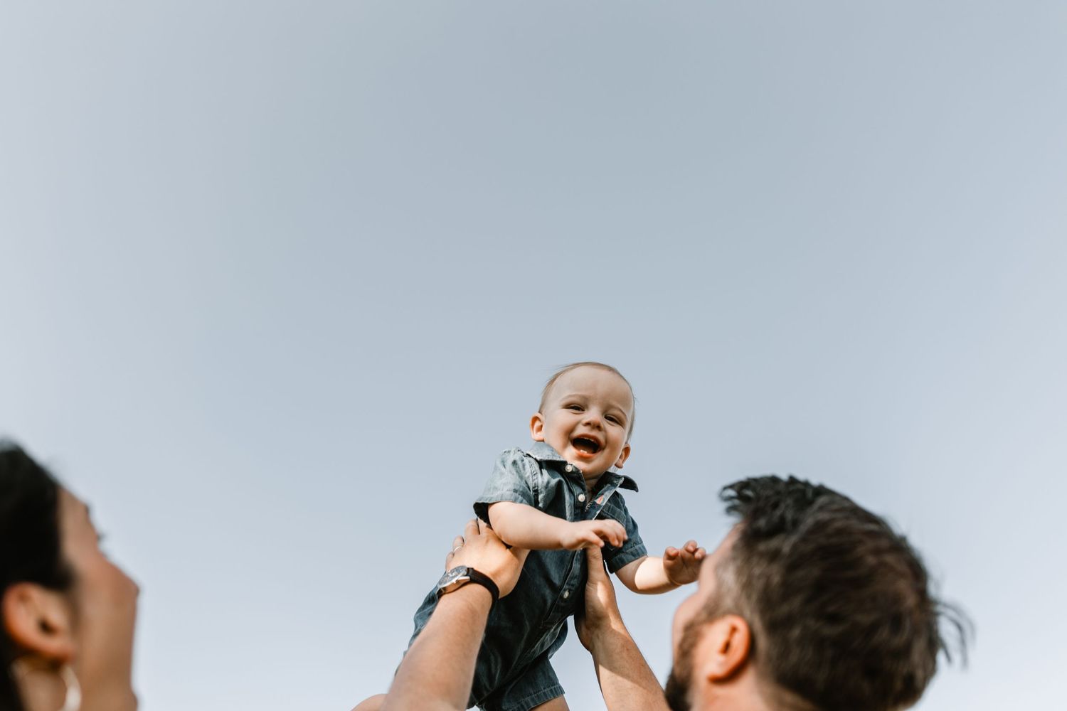 Laughing baby is lifted joyfully into the sky by parents against a clear blue background outdoors.