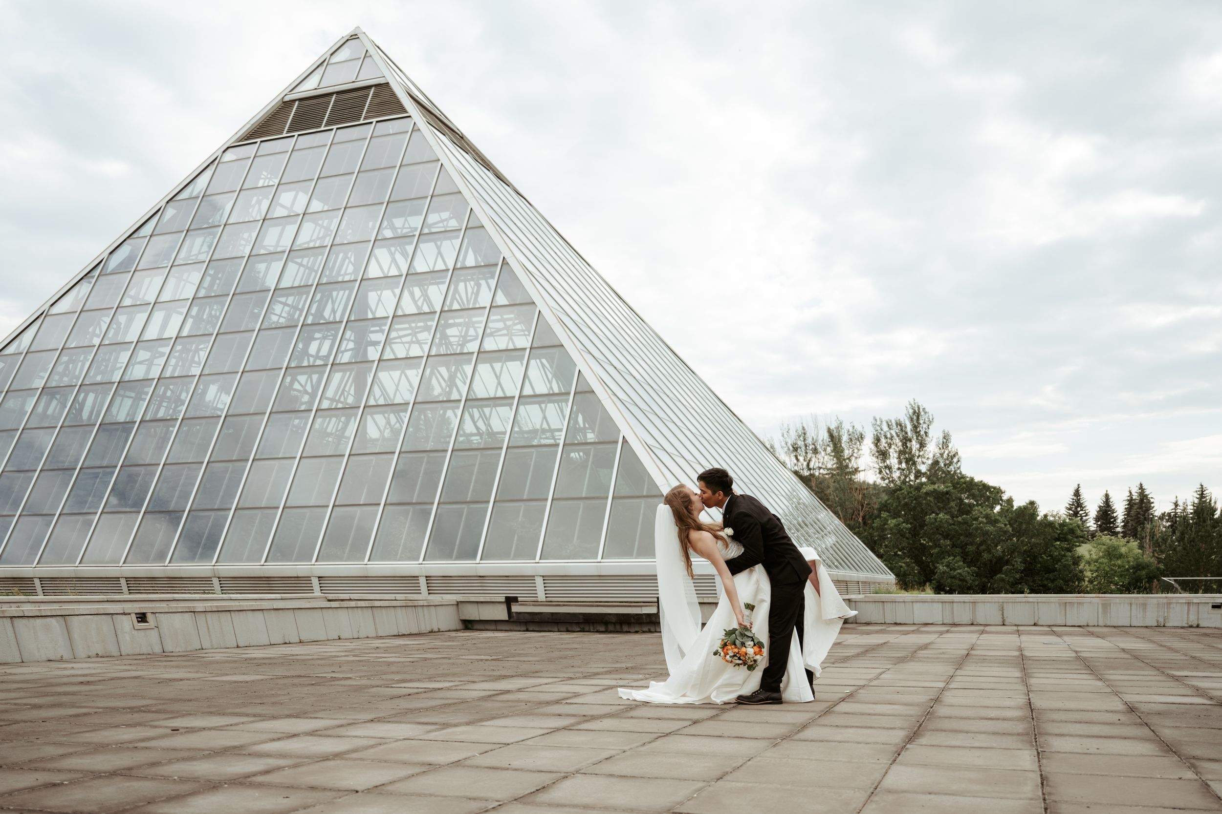 A couple embraces in front of a large glass pyramid structure under an overcast sky.