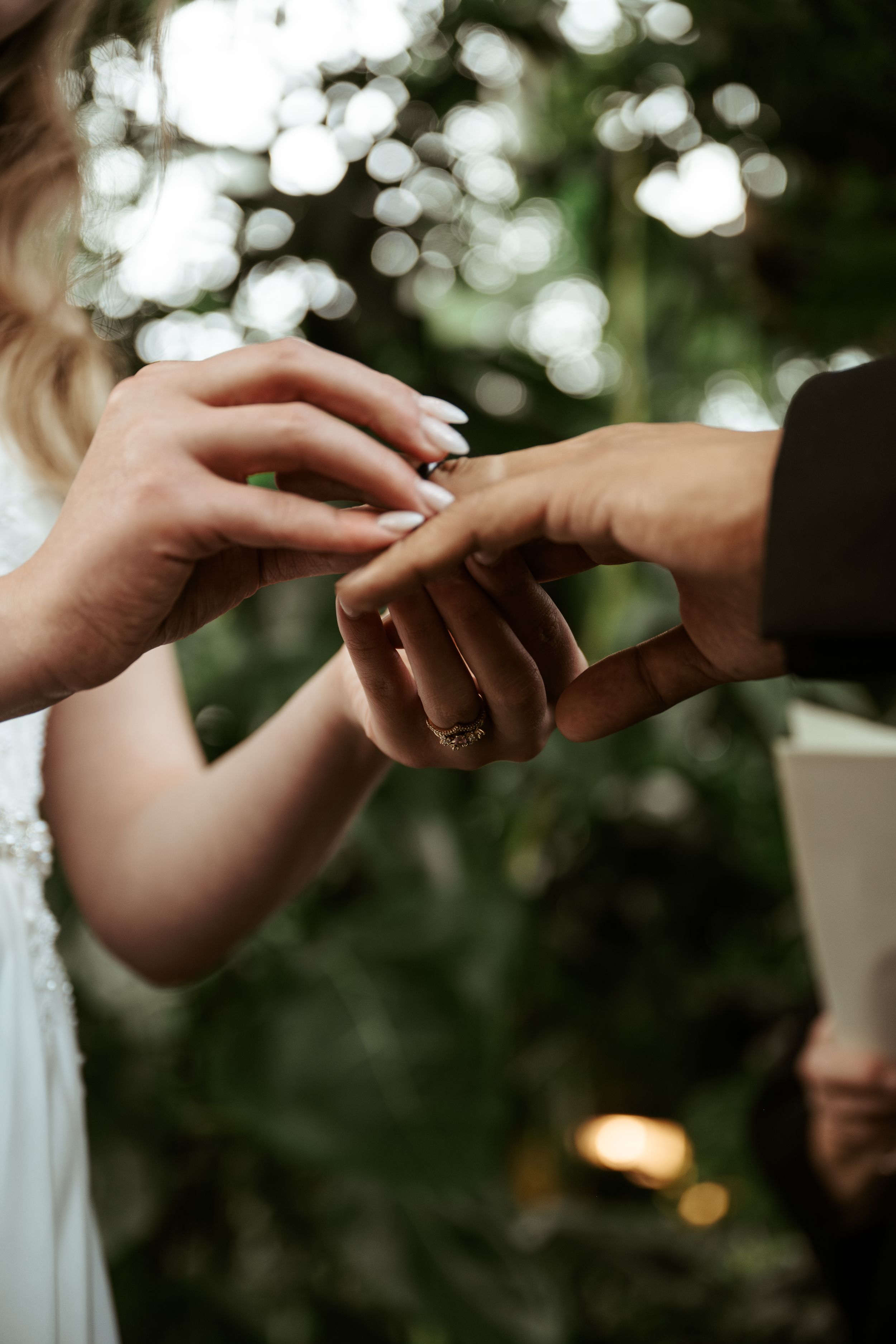 Close-up shot of hands exchanging wedding rings during an outdoor ceremony with bokeh lights in background.