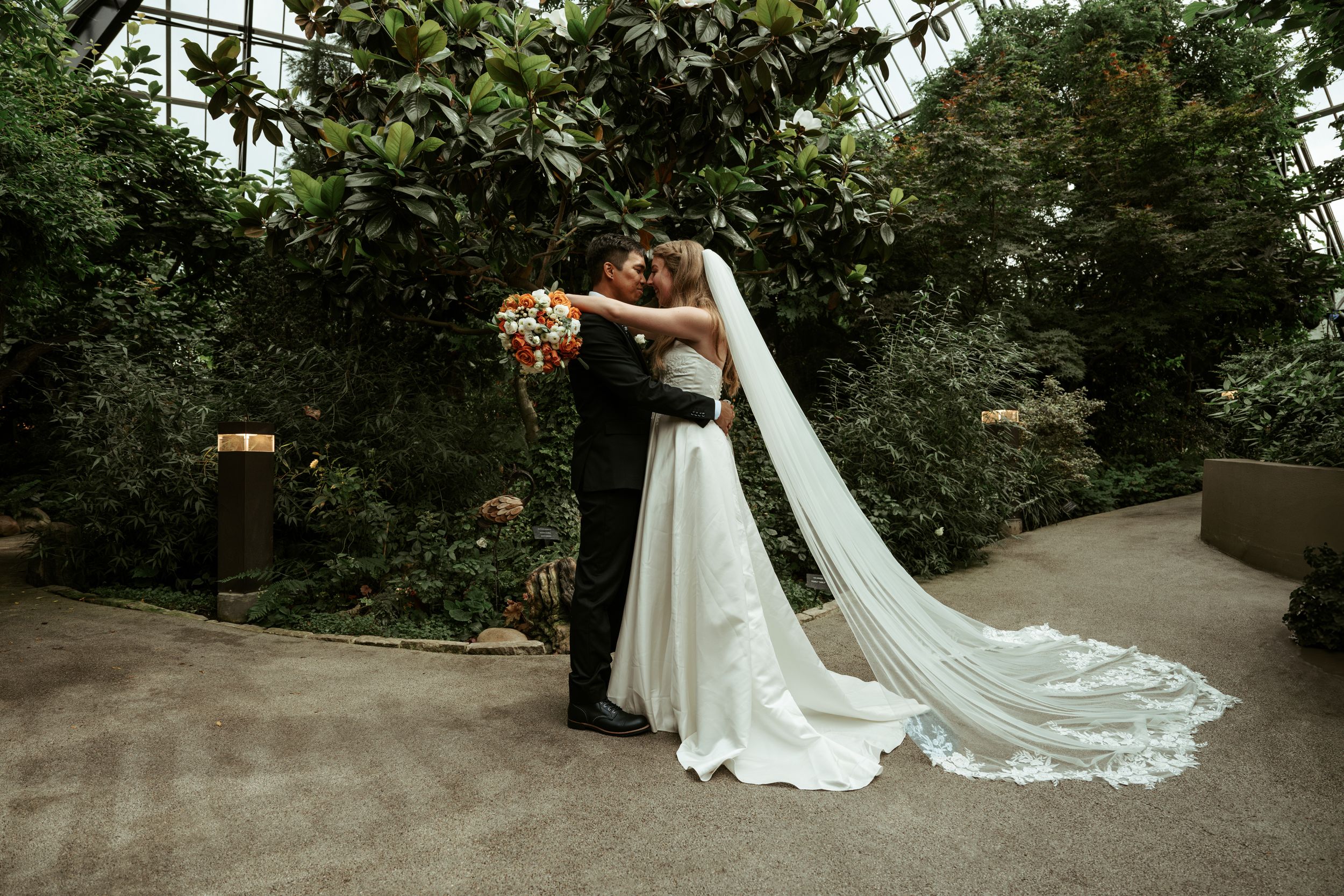 A wedding couple embraces on a garden path with a flowing white veil trailing behind them.