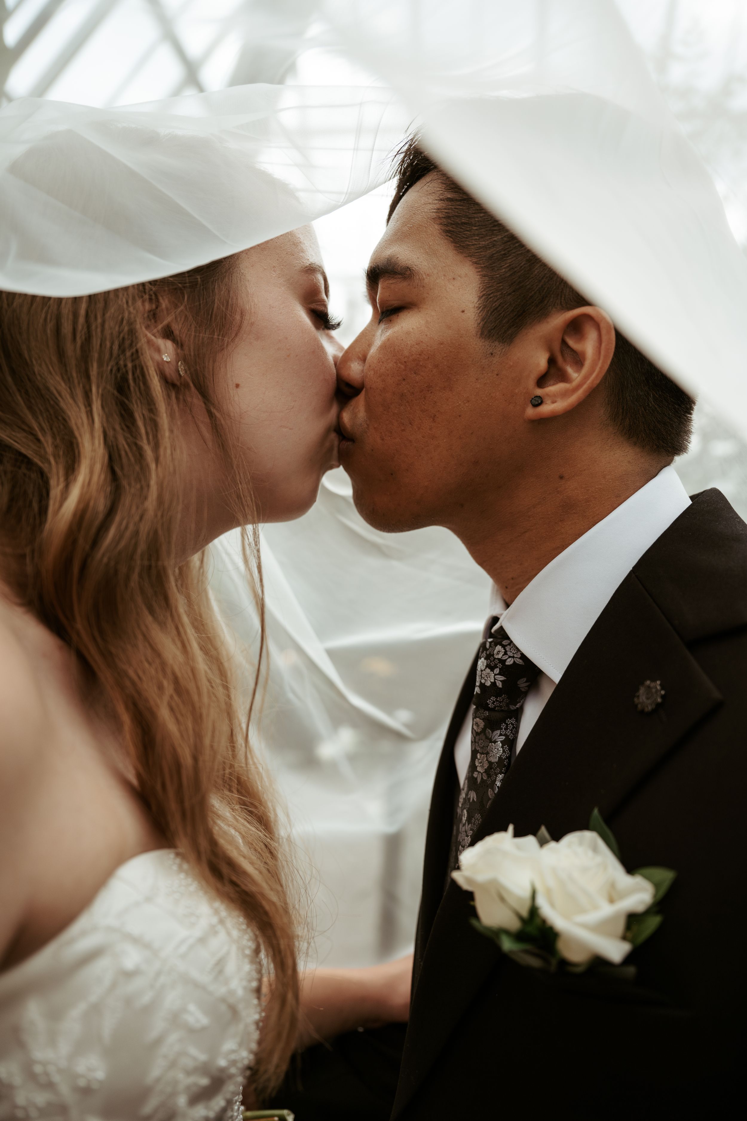 A romantic wedding kiss between a couple in traditional attire with white roses pinned to the groom's lapel.