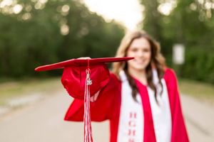 A smiling graduate in red cap and gown holds her graduation cap toward the camera on a tree-lined path.
