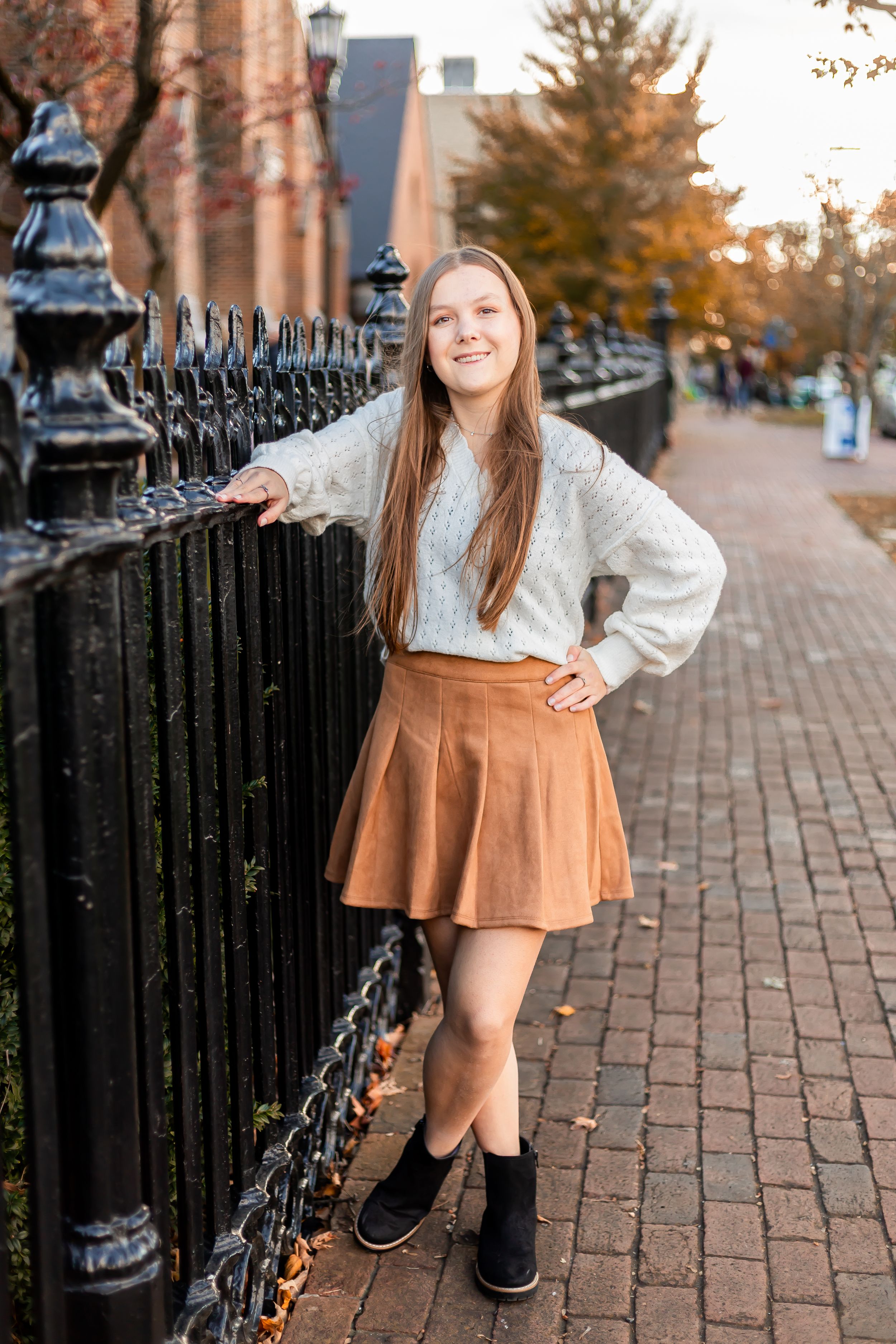 Young woman in cream sweater and brown skirt posing by black iron fence on brick sidewalk in autumn.