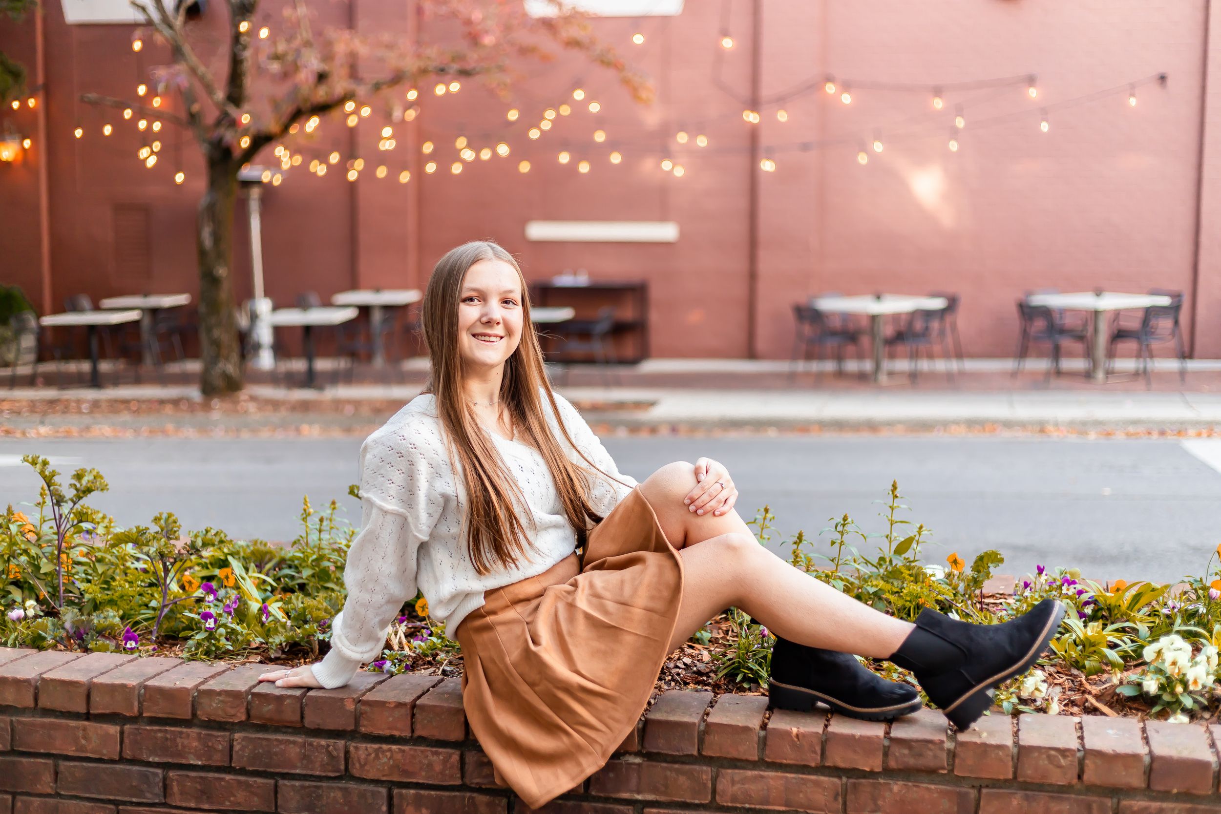 Young woman in tan skirt and white sweater sitting on brick wall outdoors with string lights and fall foliage.