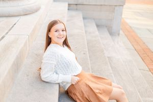 Smiling young woman in white sweater and tan skirt sitting on stone steps in warm sunlight.
