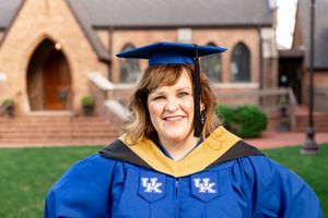 Woman in University of Kentucky blue graduation cap and gown smiling outdoors near a brick building.
