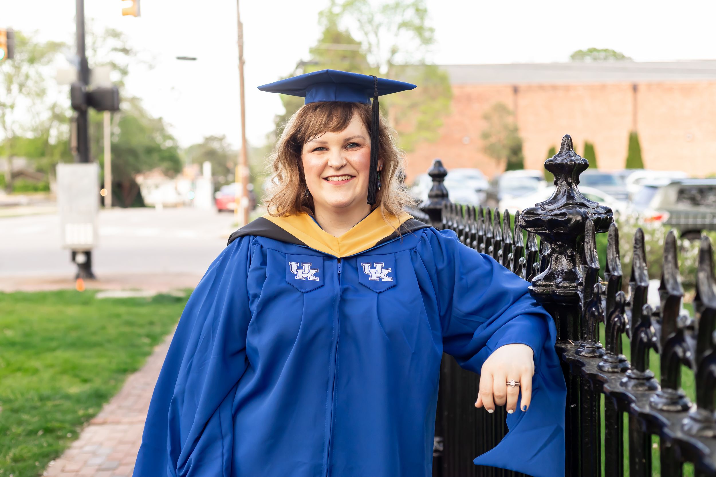 Woman in University of Kentucky blue graduation gown and cap smiling outdoors near decorative iron fence.
