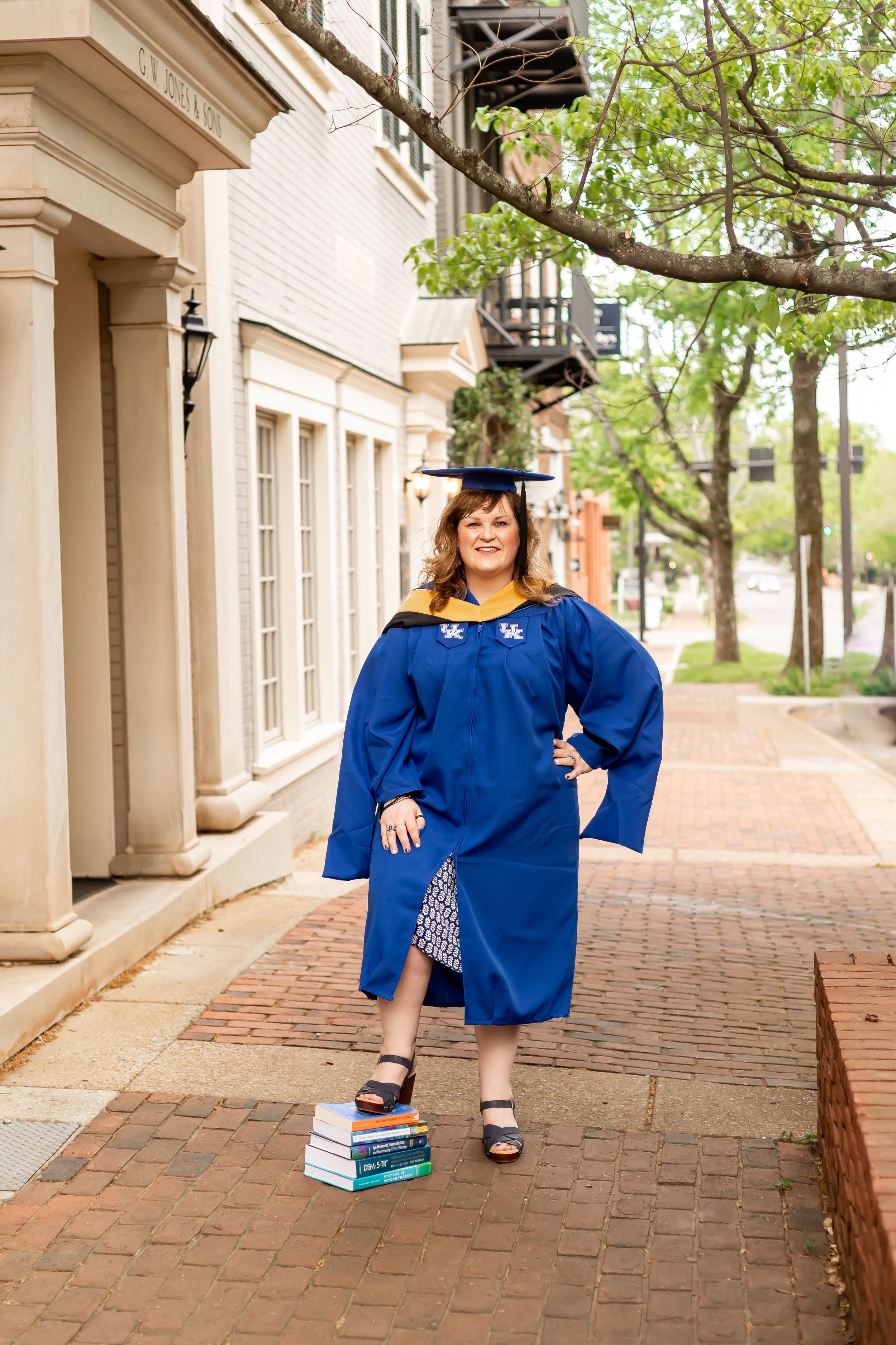 Woman in blue University of Kentucky graduation cap and gown standing on brick path with stack of books.