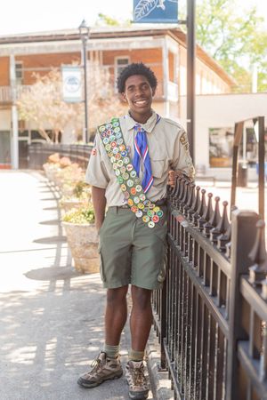 Smiling Boy Scout in uniform with merit badge sash stands by iron fence outdoors on sunny day.