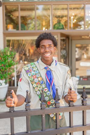 Smiling young Eagle Scout wearing merit badge sash and blue neckerchief, standing at iron fence outdoors.