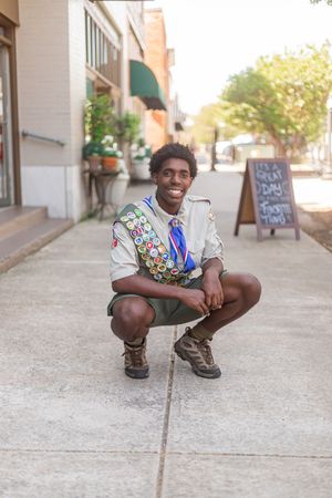 Smiling Boy Scout in uniform with merit badge sash crouching on a city sidewalk.